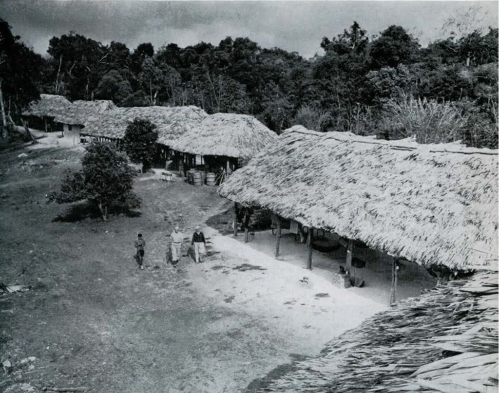View looking down on the camp, a row of thatched roof, open air buildings, three people walking by.
