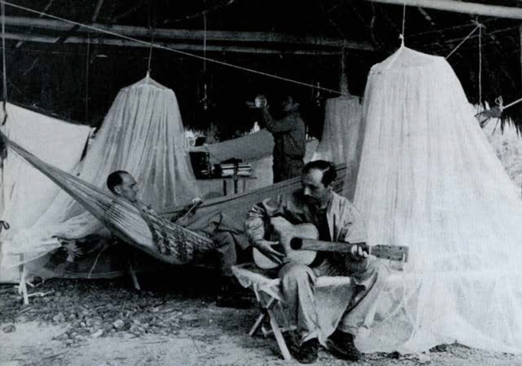 A man laying in a hammock, listening to a seated man play an acoustic guitar.