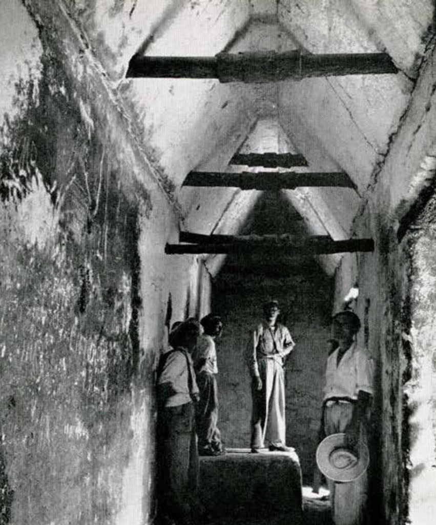 Men standing in a narrow stone hallway with vaulted ceiling and wood crossbars.