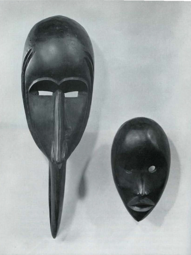 Two wooden masks mounted on a wall, left mask has beak of a bird in place of nose.