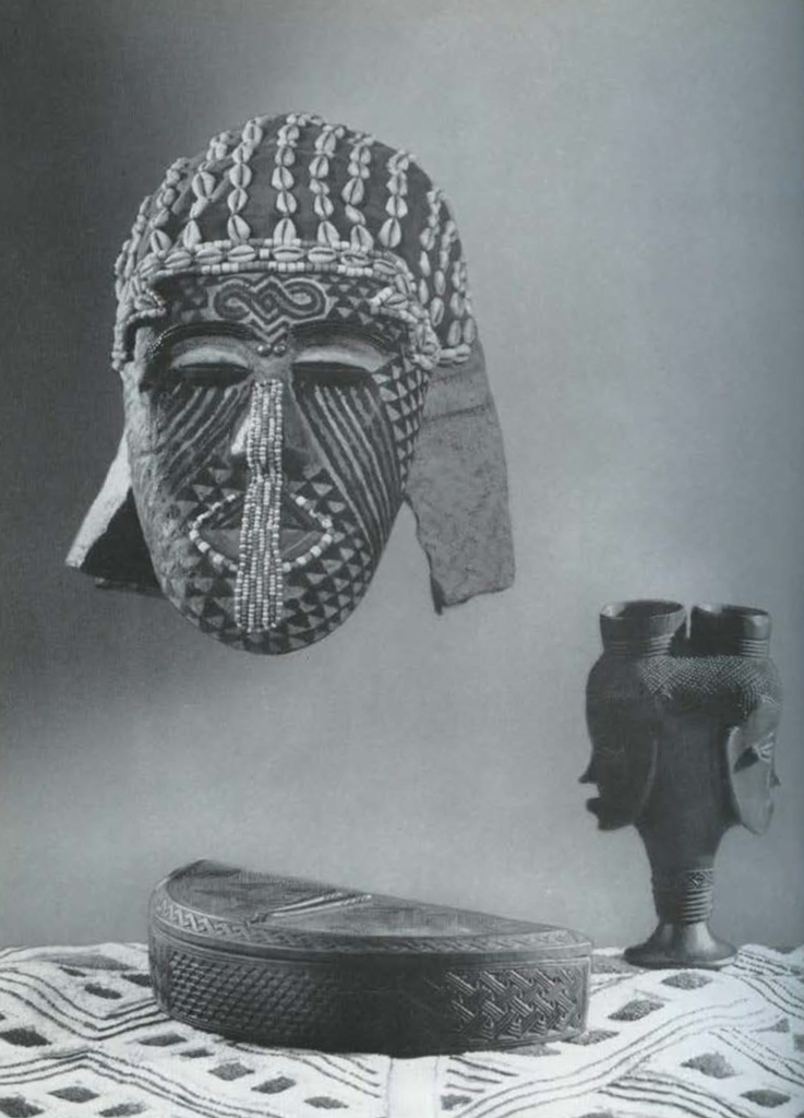 A beaded mask, half circle box, wooden cup of two faces, and rafia cloth with geometric pattern.