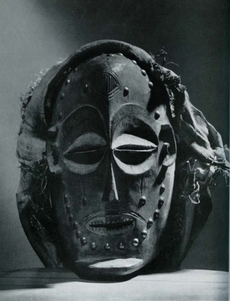 Wooden mask with slits for eyes and mouth; sharp pointed teeth; prominent chin; incised lines (scarification?) on forehead and upper lip; studded with brass-headed nails, to top of mask is attached a cap of palm cloth over a framework of twigs.