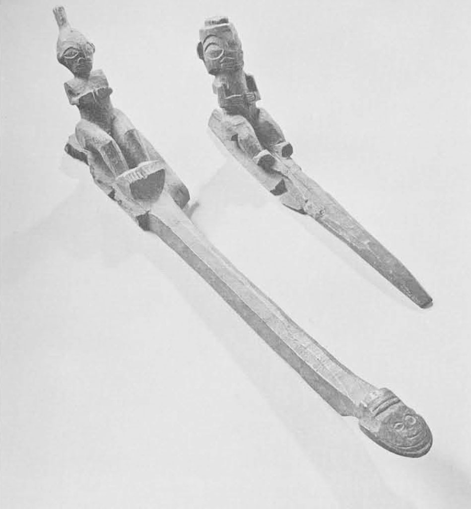Two carved wooden canoe ornaments, featuring seated figures leaned back as if rowing at the end of each.