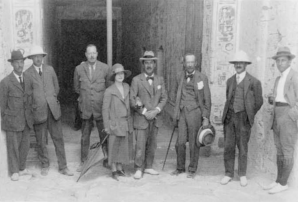 A group photo of seven men in suits and one woman in a long coat, standing outside painted tomb walls.
