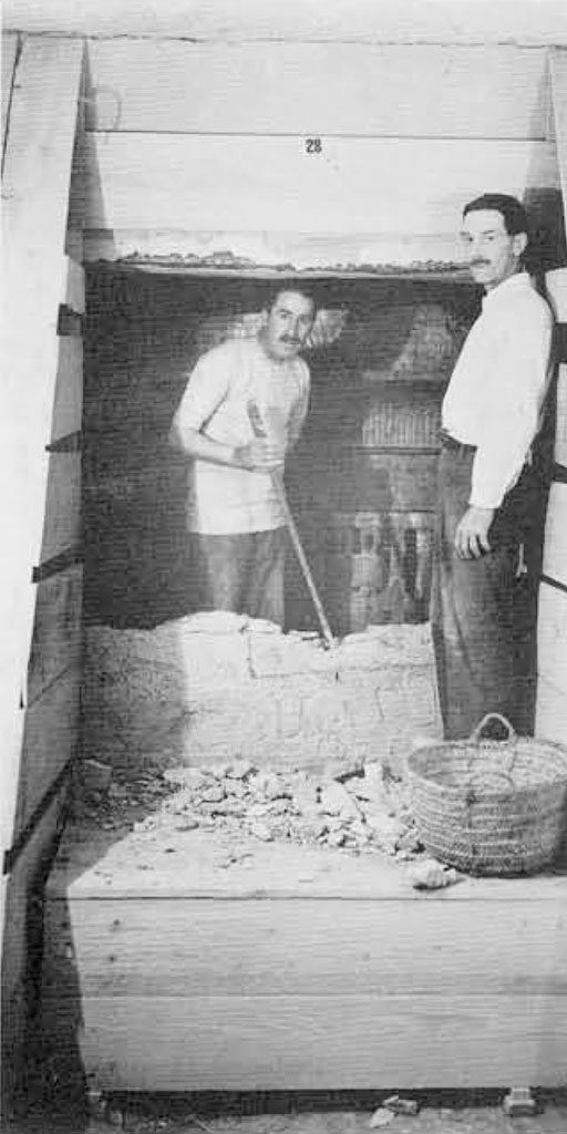 Two men in a tomb, walls propped up with wood, using a crowbar to open a door.