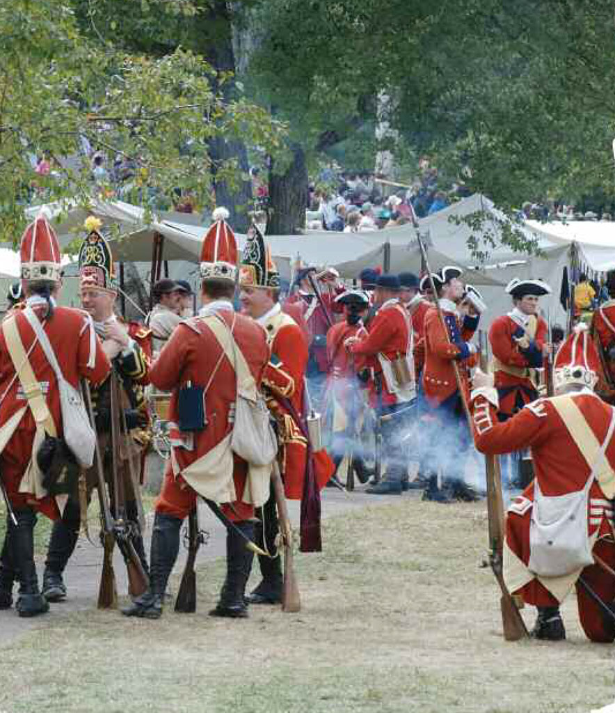 A field full of re-enactors in period clothing, standing in front of white tents and holding guns.