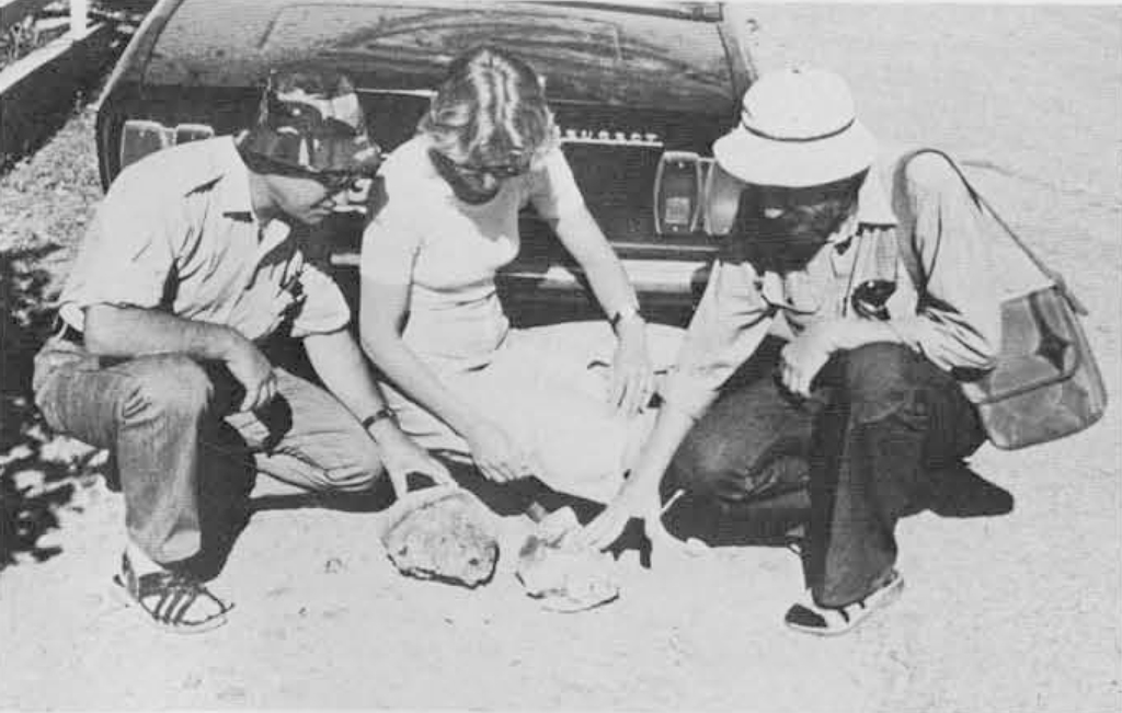 Three archaeoligists crouching on the ground in front of a car, touching two objects in front of them.