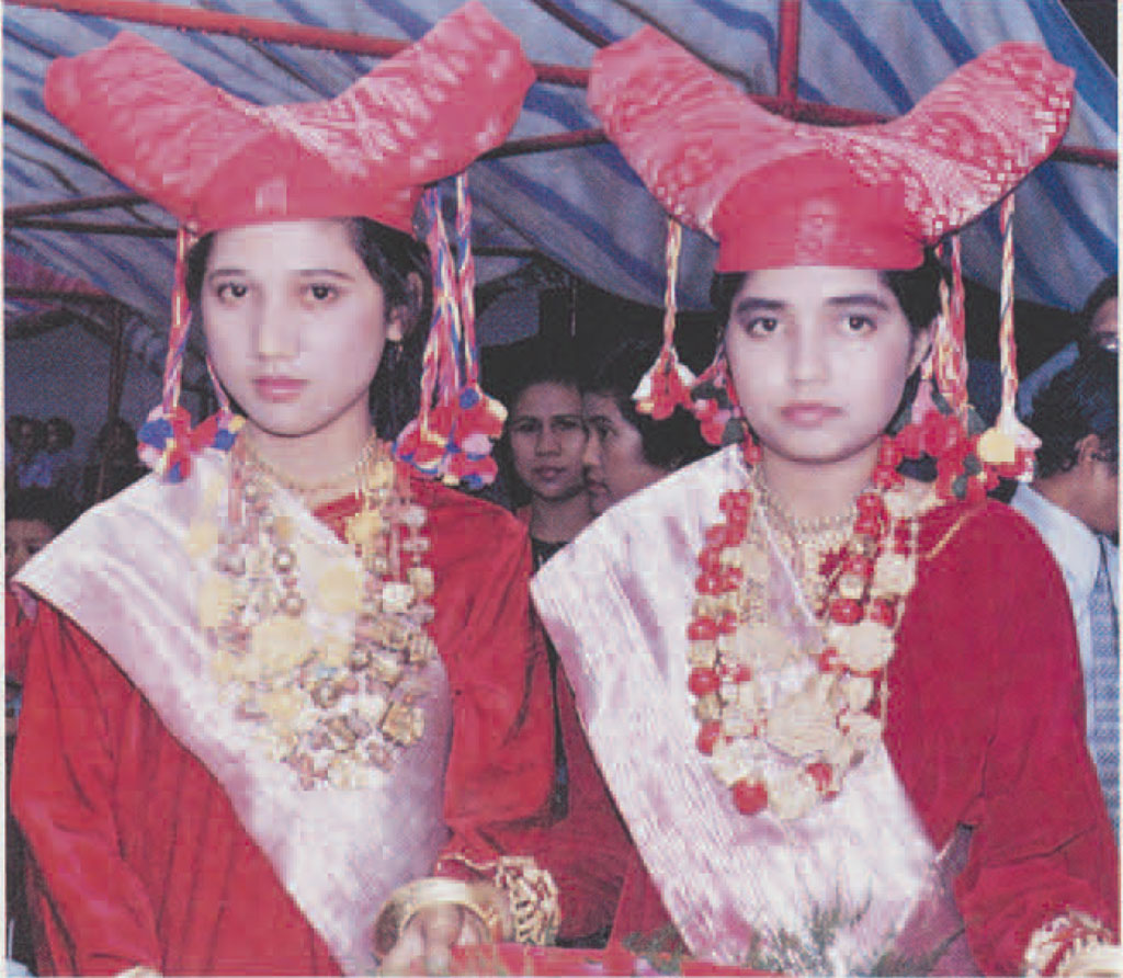 Two women in identical clothes, a red tunic with wide pink sash, flower necklaces, and pronged red headdress with tassels.