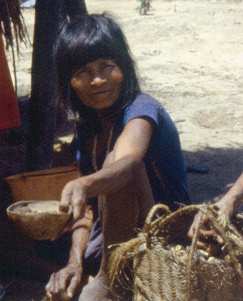 A woman sitting next to a basket of roots, holding out a bowl of food.