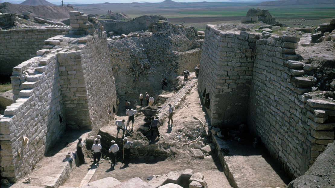 A large corridor or street at Gordion with high stone walls surrounding, a group of workers putting debris on carts on a track in the corridor.
