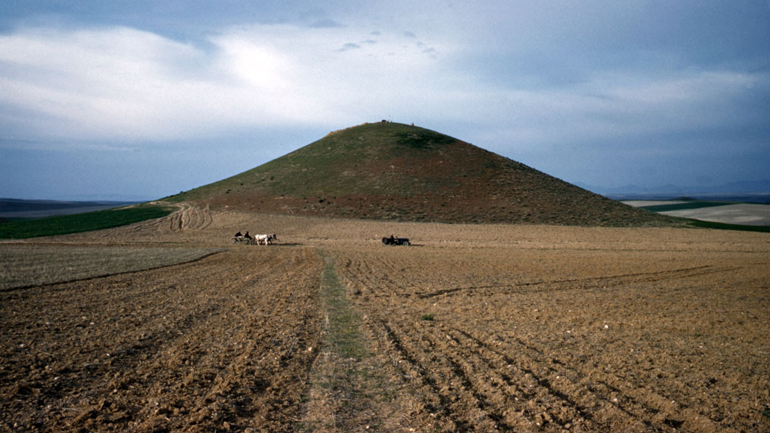 A plowed field with a massive mound in the distance. A horse and buggy cross the field.