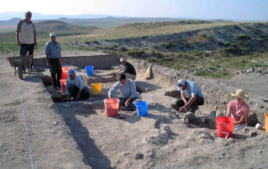 A group of people. crouching in a rectangular depression, working to excavate artifacts.