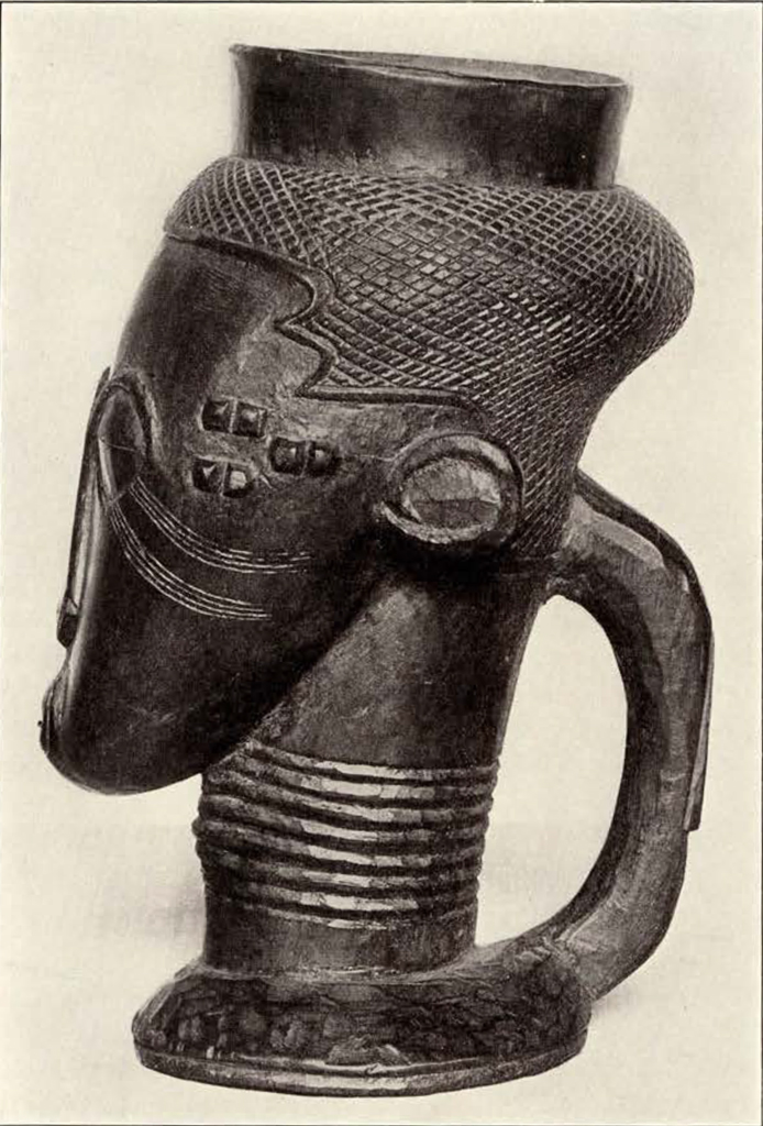 A cup in the shape of a head and face with long neck, from the side showing cup handle and rings on the neck