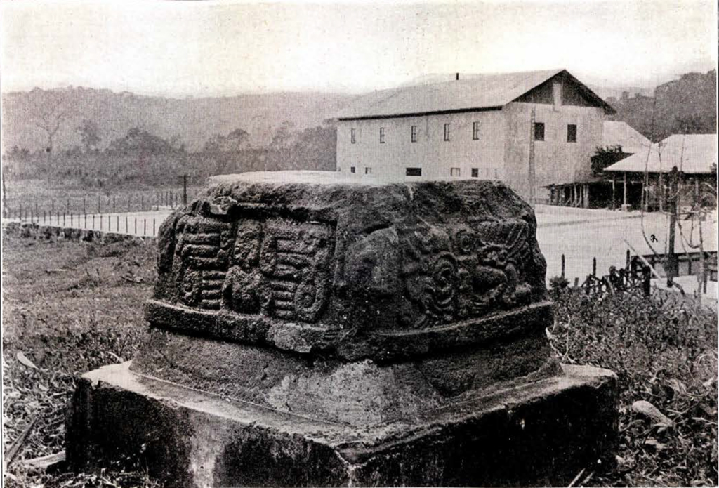A stone with designs carved into the sides on a square dias, a building behind and mountains in the distance