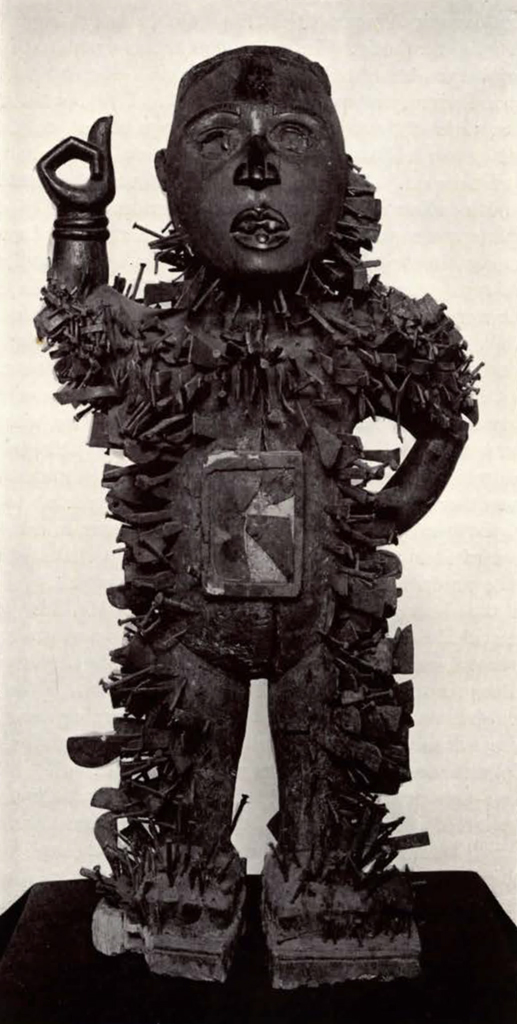 Wood carved figure, standing, with one arm on hip and one arm in the air, covered in holes, many empty, many with nails driven in