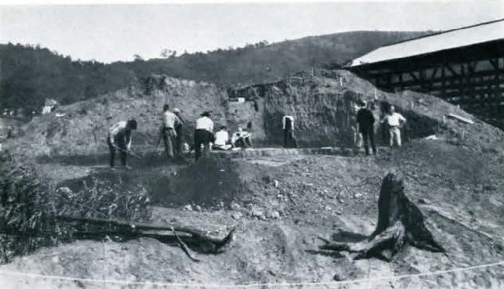 A mound being excavated, several people digging a huge pit
