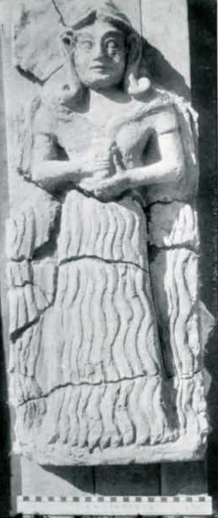 Relief of a goddess with hair curled at the ends, wearing an off the shoulder long dress with hands holding a vessel at the waist