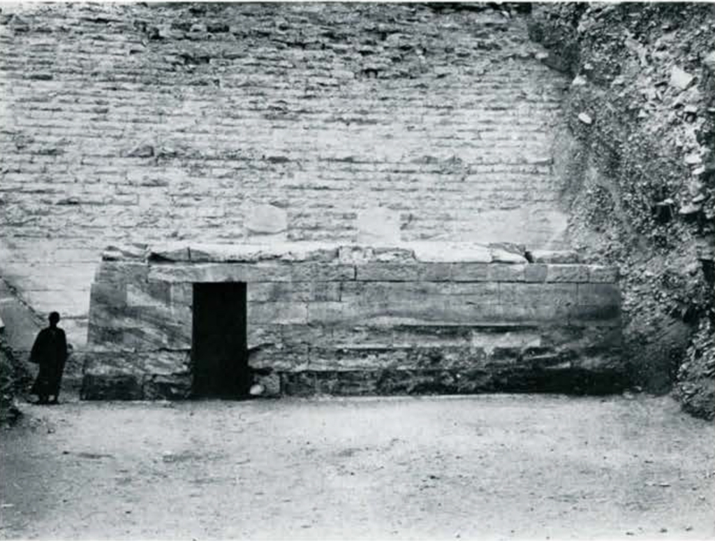 Low brick building with a entrance in front of or within the pyramid from the front with two stelae in the back stick up over the roof