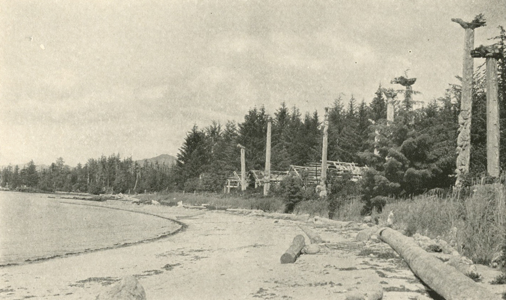 A beach with shrubbery and trees, a few small houses or huts and totem poles lining it