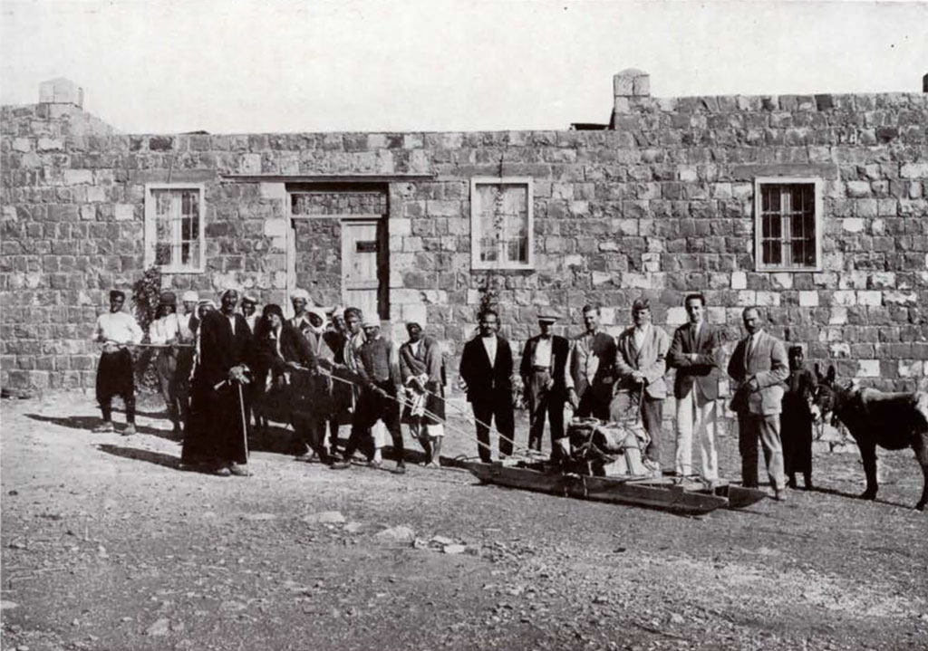 A large group of men standing in front of a brick house, a column base on a sled being bulled