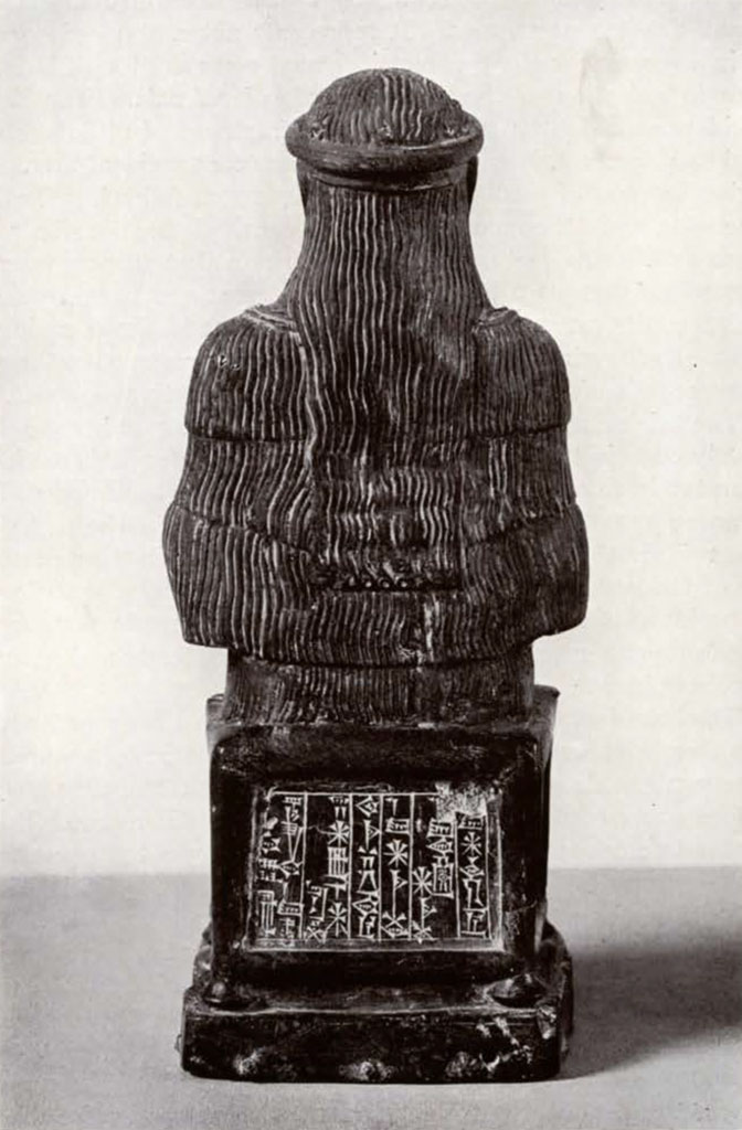 Seated goddess wearing tiered dress with headband, back view showing long hair and inscription